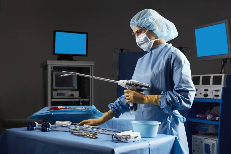 mulher com roupa cirúrgica manuseia equipamento em sala com computadores e monitores, há uma mesa à sua frente com instrumentos cirúrgicos