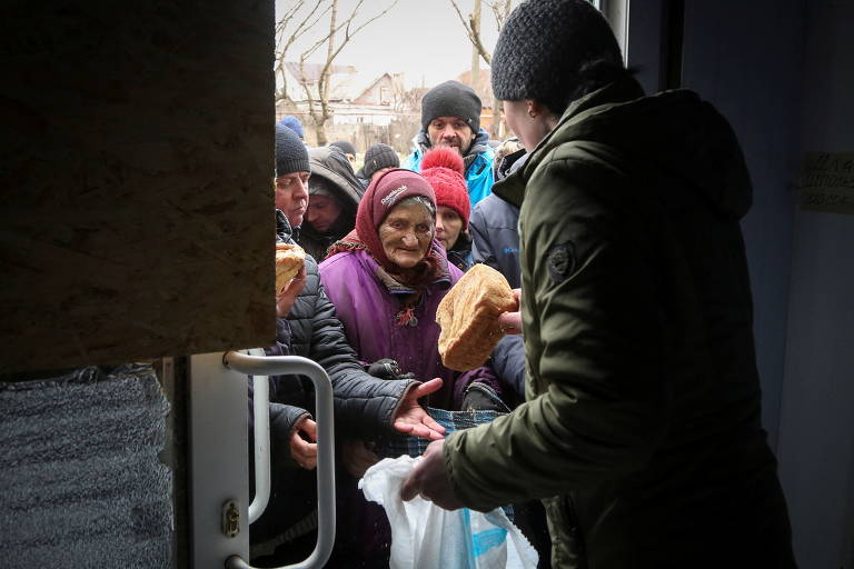 Voluntário entrega pão para uma mulher idosa, que cobre a cabeça com um lenço 