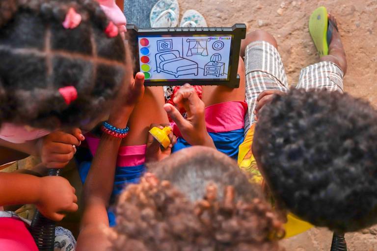 Imagem colorida, tirada de cima, mostra três crianças manuseando um tablet