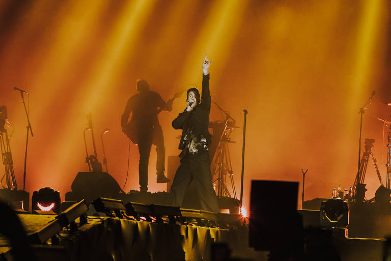 Fotografia colorida do palco do show do Maroon 5; vocalista Adam Levine está ao centro com um microfone e a mão levantada em um cenário com luz amarela