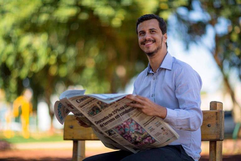 Imagem em primeiro plano mostra homem jovem sentado em um banco de praça. Ele está sorrindo e com um jornal na mão.