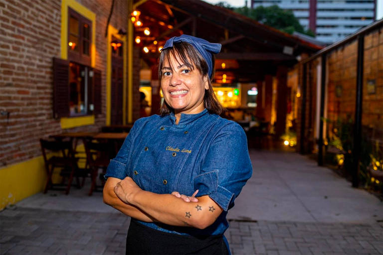 A foto mostra a chef Claudinha Luna de dolman azul escuro com o restaurante ao fundo.