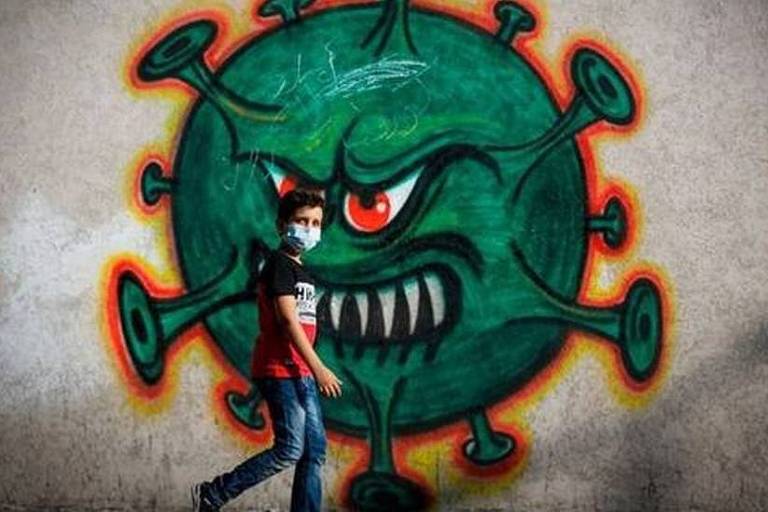 Imagem panorâmica mostra um menino de máscara caminhando. Ao fundo, em uma parede, há a ilustração de um coronavírus com olhos e boca e semblante raivoso.