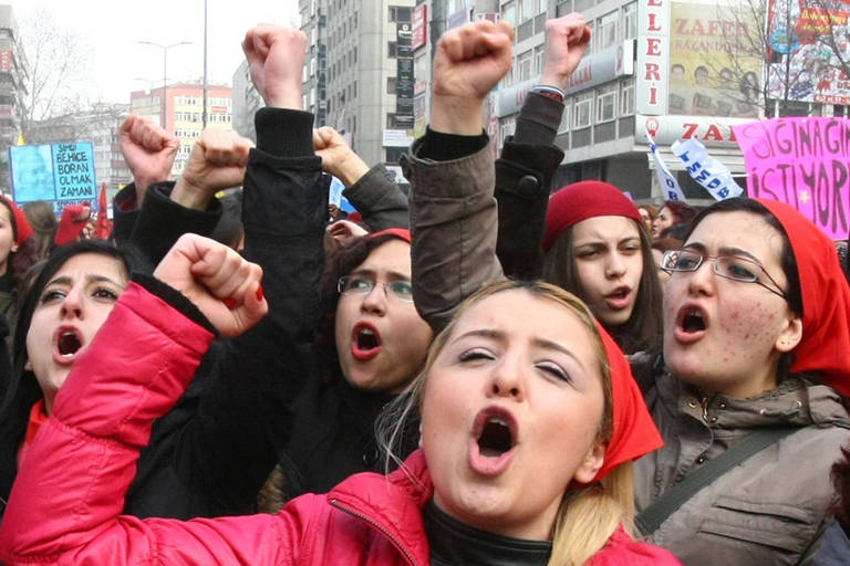 Mulheres gritam, erguendo os punhos para o ar em protesto; elas cobrem o cabelo com um pano vermelho.