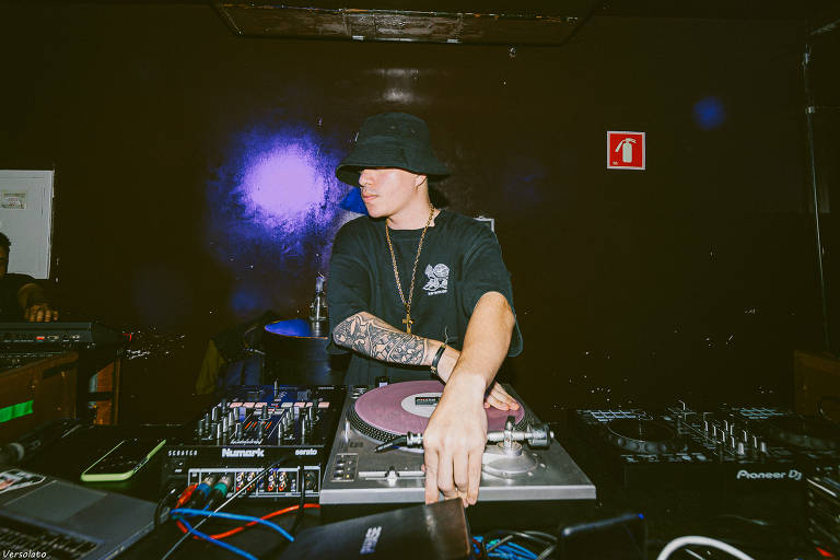 DJ branco com chapéu preto e camisa preta. Em baixo das mãos está um toca disco