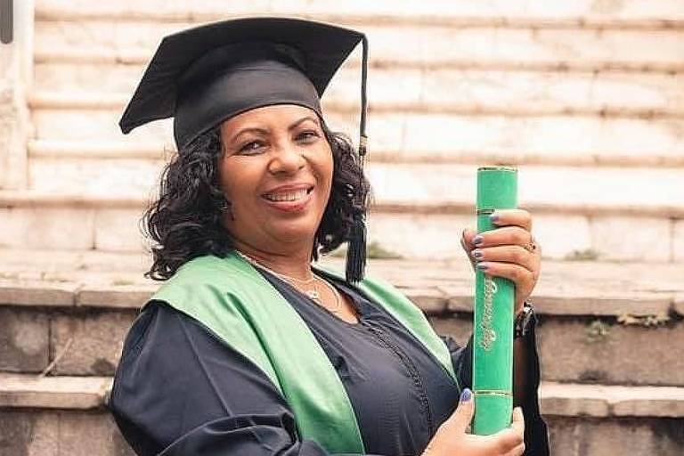 mulher de meia idade, de pele parda, vestida com beca de formatura e chapéu característicos pretos mostra seu canudo verde contendo deu diploma
