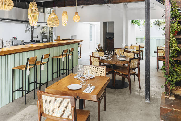 Ambiente interno do restaurante, bem iluminado, com mesas e cadeiras de de madeira e balcão verde.