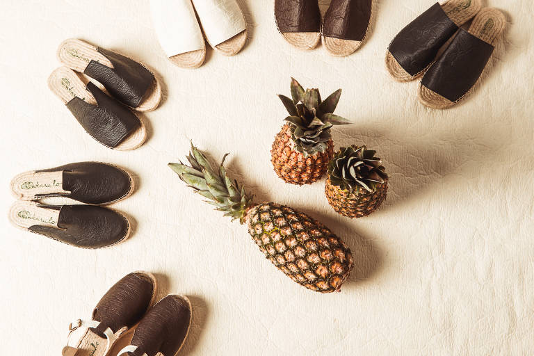 Seis pares de sapato aparecem em semicírculo sob um tapete bege. No centro da imagem está um abacaxi, indicando a relação do material dos calçados com a fruta
