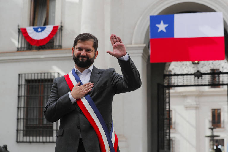 Vestido com a faixa presidencial, o presidente do Chile, Gabriel Boric, que tem barba preta, está com a mão direita no peito e a esquerda erguida no Palácio de La Moneda, em Santiago; atrás dele aparece a bandeira chilena