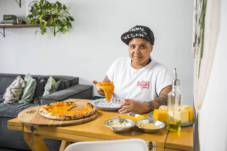 Isabela está sentada em uma mesa segurando um pedaço da pizza vegana. Ela é uma mulher negra que veste boné preto escrito "vegan" e camisa branca. 