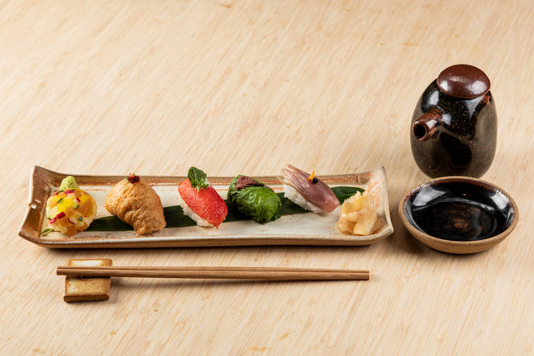 Prato retangular com cinco versões de sushis vegetarianos do restaurante Aizomê; ao lado do prato estão hashis e cerâmicas de shoyu