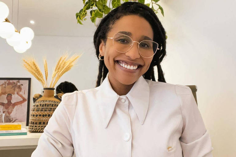 Fotografia colorida em plano médio de uma mulher negra que sorri e veste um jaleco branco em uma das salas da sua clínica médica chamada Derma Vegan, Faria Lima, zona sul, SP. 