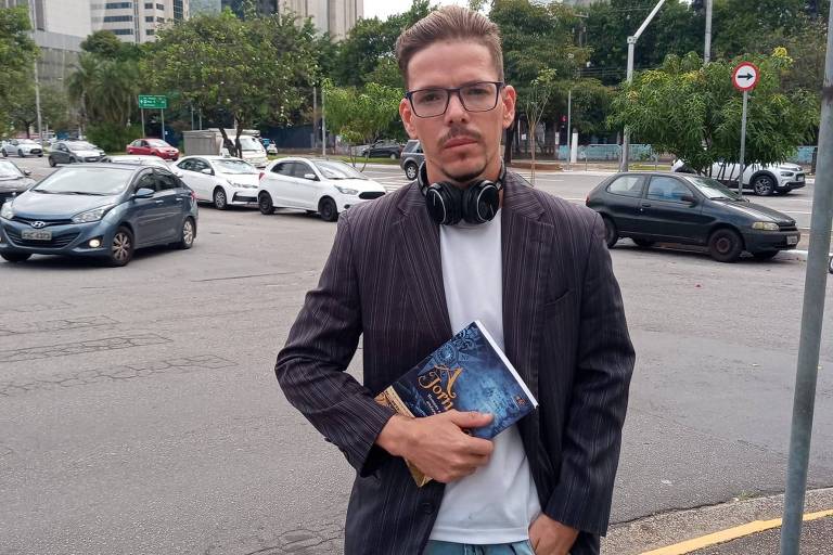 Gerson Lavisio usa óculos, veste um paletó sobre uma camiseta. Ele está com um fone de ouvido preso ao pescoço e segura um livro em uma das mãos