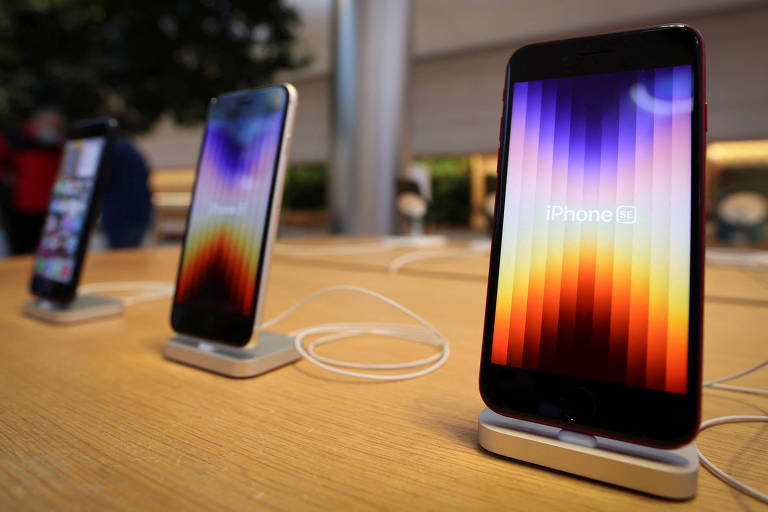 Três smartphones com a tela colorida estão expostos na vertical sobre uma mesa de madeira.