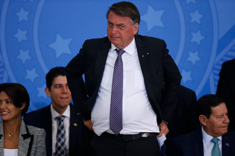 Em primeiro plano, Jair Bolsonaro está em pé, e faz um gesto de levantar as calças com as duas as mãos; usa terno e tem uma expressão de desconforto; ao fundo, sua esposa e dois membros do governo estão sentados em cadeiras