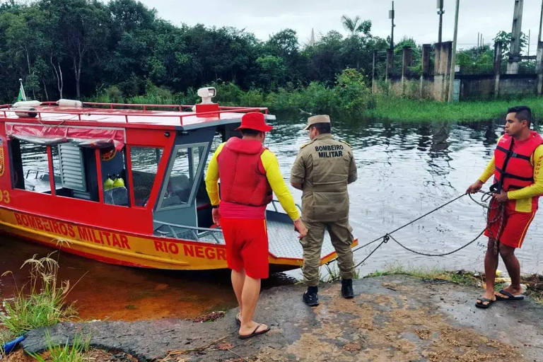 Homens com coletes salva-vidas vermelhos puxam com corda uma embarcação também vermelha na beira de um rio