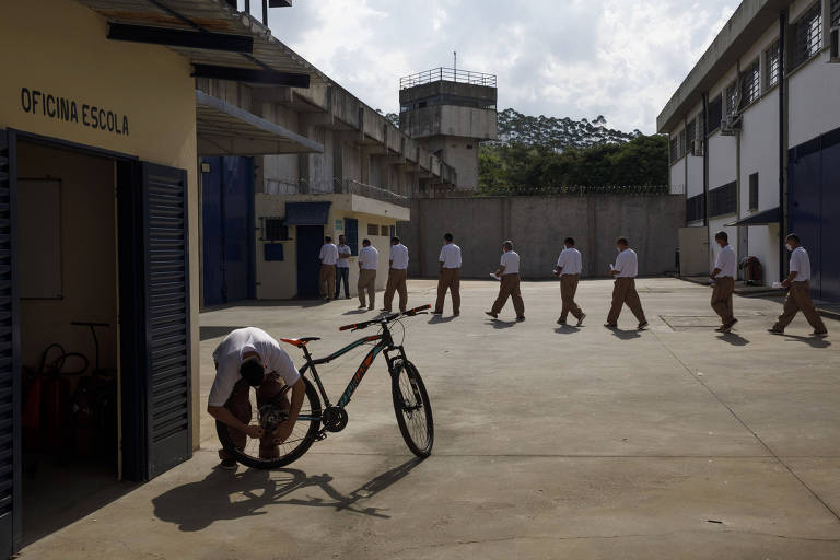 atrás de homem que se agacha para consertar bicicleta, passa uma fila de presos