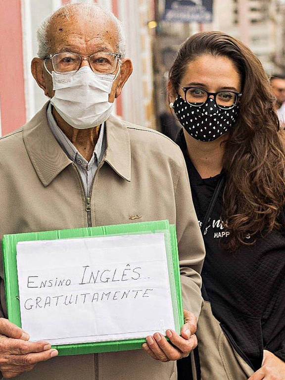 homem de máscara idoso de óculos ao lado de jovem com cabelos compridos castanhos de máscara. ele segura cartaz: reforço de inglês, aulas gratuitas