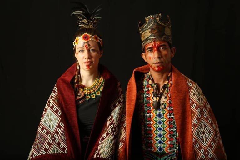 Mulher branca e homem indígena, em trajes étnicos, posam lado a lado