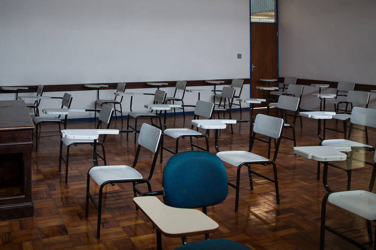 Foto mostra sala de aula vazia, cadeiras brancas estão espalhadas pelo espaço de chão de madeira. Ao fundo pode-se observar uma lousa branca, sem nada escrito