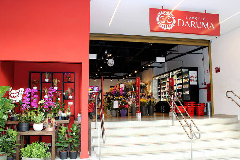 A foto mostra a fachada de uma loja com uma parede vermelha com vasos de flores e ao lado, uma escada de acesso à loja