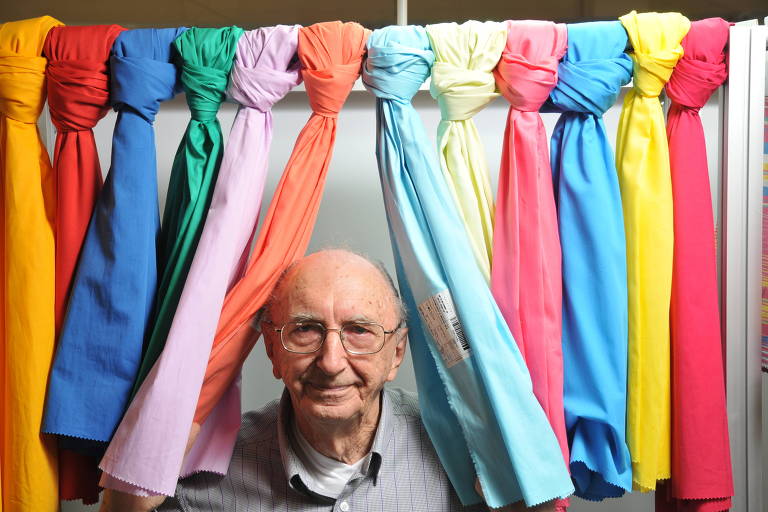 homem idoso posa para a foto em meio a panos coloridos amarrados em haste acima dele