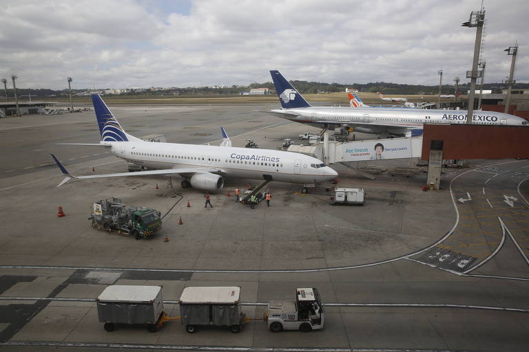 Imagem mostra pista de decolagem e pouso de um aeroporto. É possível ver dois aviões na zona de embarque. Os dois são brancos, com a cor azul na parte traseira. Há funcionários e carrinhos com bagagens na pista.
