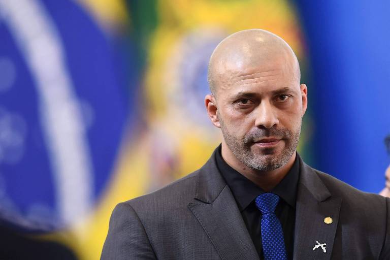 O deputado Daniel Silveira veste terno e camisa escuros com gravata azul. Ao fundo, pode-se ver bandeiras do Brasil.