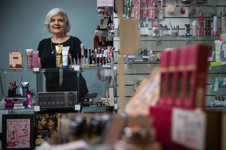 Mulher de cabelos brancos e roupa preta está atrás de um balcão de vidro repleto de cosméticos e maquiagens