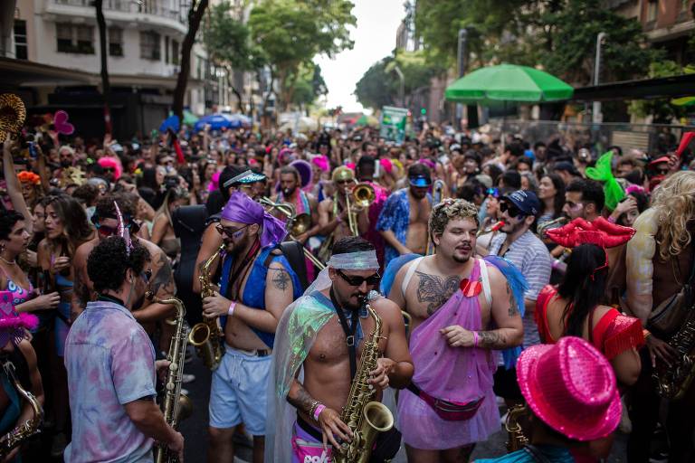 multidão em bloco de rua com fantasias em uma rua do Rio; à frente um homem fantasiado toca saxofone
