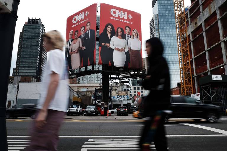 Fotografia mostra uma avenida movimentada; duas pessoas caminham em primeiro plano, em segundo, está um anúncio vermelho em um topo de um prédio com o logo 'CNN plus'