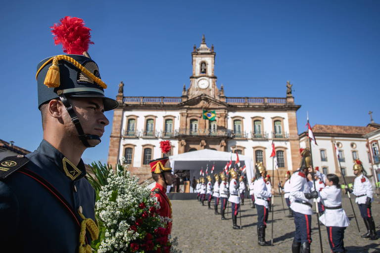 Guarda uniformizado segurando flores é visto em primeiro plano; ao fundo oficiais enfileirados na Praça Tiradentes, construção em estilo colonial e céu azul 