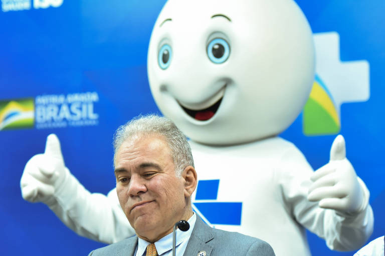 Marcelo Queiroga sorri com a boca repuxada, enquanto, atrás dele, o personagem Zé Gotinha faz sinal de positivo com as duas mãos