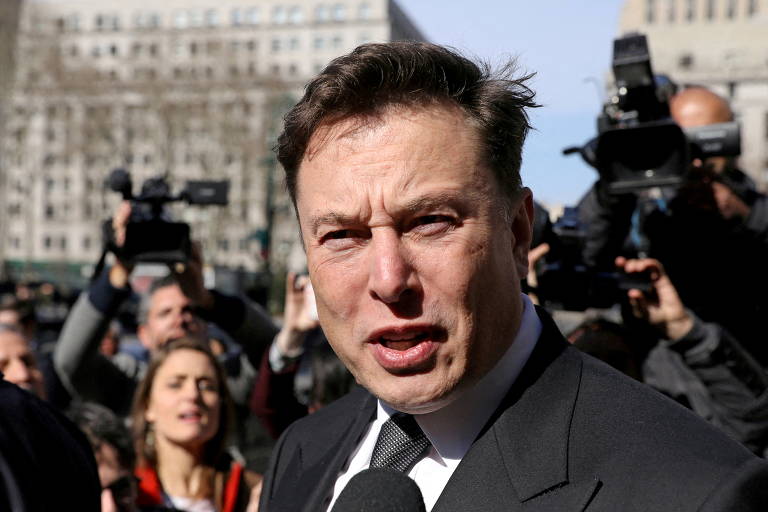 Elon Musk, um homem branco e de cabelos negros, aparece na foto dando entrevista para repórteres. Ele veste terno preto, camisa social branca e gravata preta. Há um microfone à sua frente e, ao fundo, há vários repórteres e pessoas com câmeras
