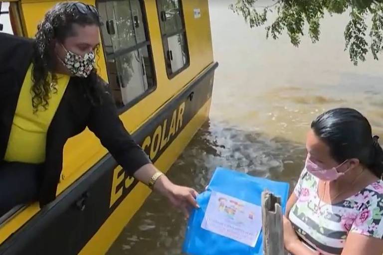 Mulher em barco entrega materiais escolates a mulher na beira do rio