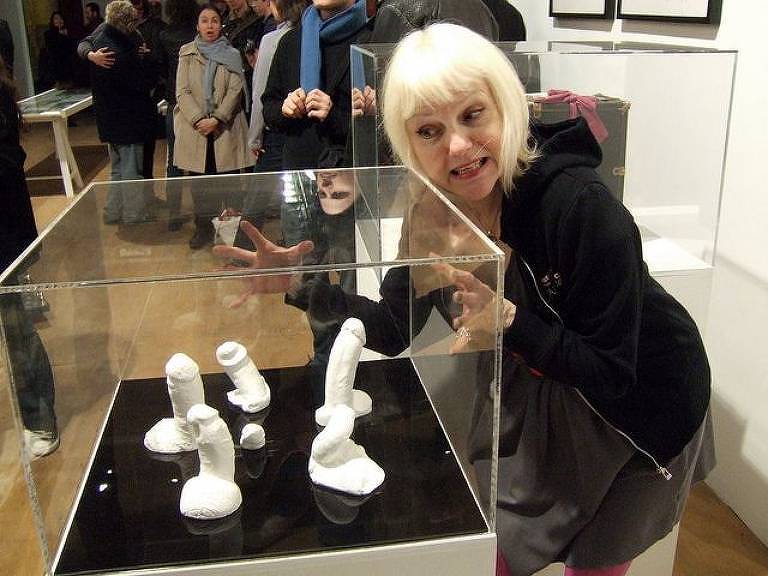 A artista Cynthia Albritton, conhecida como Cynthia Plaster Caster, ao lado de pênis de gesso em exposição