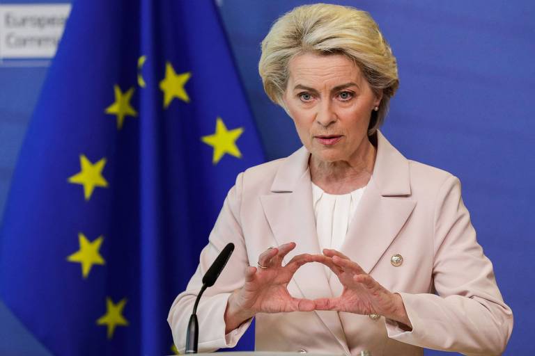 Mulher em terno creme fala frente a bandeira a União Europeia