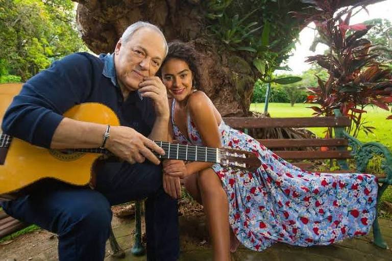 Em foto colorida, o violonista, compositor e cantor Toquinho e a cantora Camilla Faustino posam para a câmera sentados em um banco de jardim
