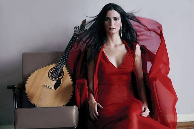 Em foto colorida a instrumentista Marta Pereira da Costa posa para a câmera usando vestes vermelhas ao lado de uma guitarra portuguesa
