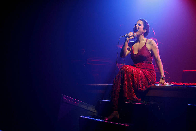 Em foto colorida, a cantora Verônica Ferriani aparece cantando sentanda em um piano e trajando vestido vermelho