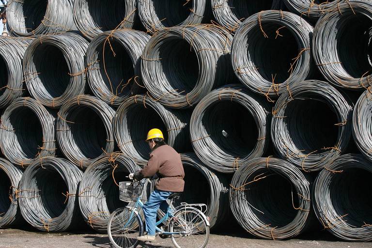 Trabalhador passa por bobinas de metal em área de armazenamento de ferro e aço no noroeste da China
