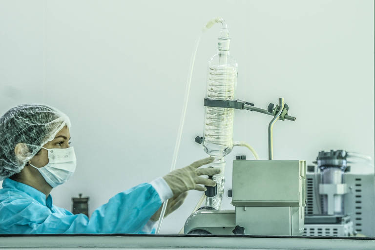 Imagem colorida mostra mulher, do ombro para cima, voltada para o lado direito. Ela usa touca transparente na cabeça, avental azul e luvas brancas. Está manuseando um aparelho dentro de um laboratório.