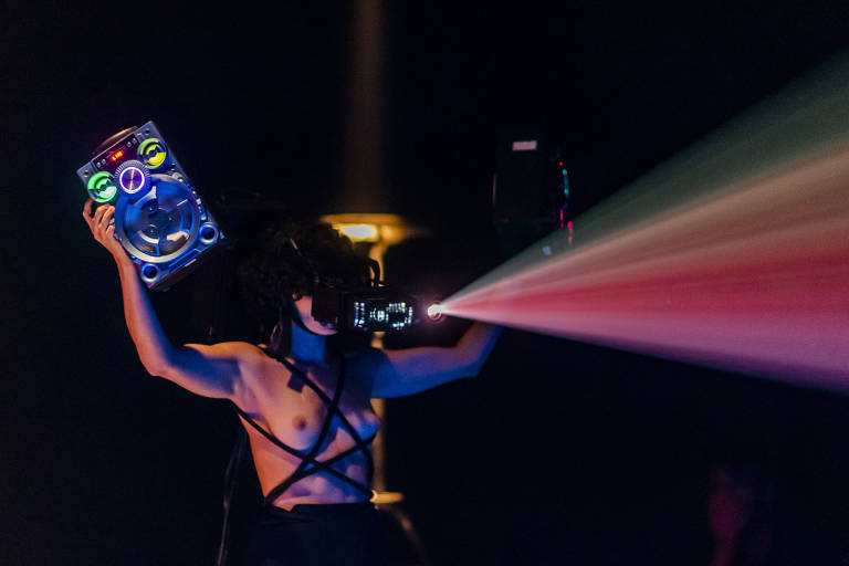 Um corpo feminino segura equipamentos que emitem luzes coloridas neon num palco
