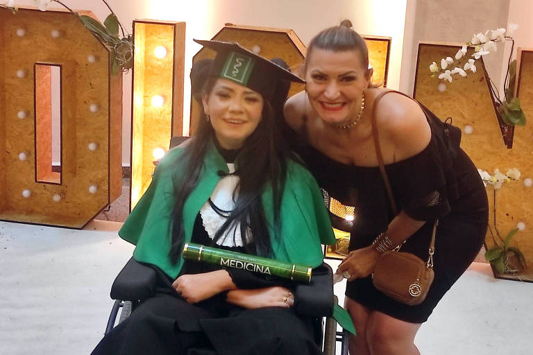 mulher de cadeira de rodas com roupa de formatura e canudo escrito medicina, na cor verde, com mulher de pé ao lado