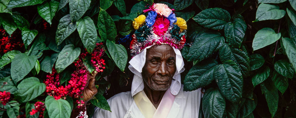 homem negro com roupas folclóricas posa apoiado em parede viva, cercado por plantas