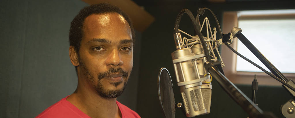 Homem negro vestindo camiseta vermelha em estúdio de gravação de frente para um microfone
