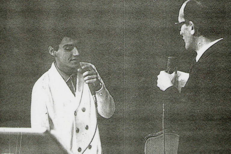 foto em preto e branco mostra rapaz de terno brnaco com microfone cantando em frente a homem de terno escuro