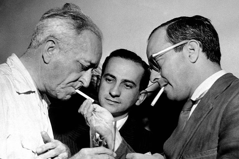 O compositor e poeta Jamie Ovalle, à esquerda, com Otto Lara Resende, ao centro, e Vinicius de Moraes, à direita, em 1953. Vinicius de Moraes, de terno e óculos escuros, acende o cigarro de Jamie Ovalle.
