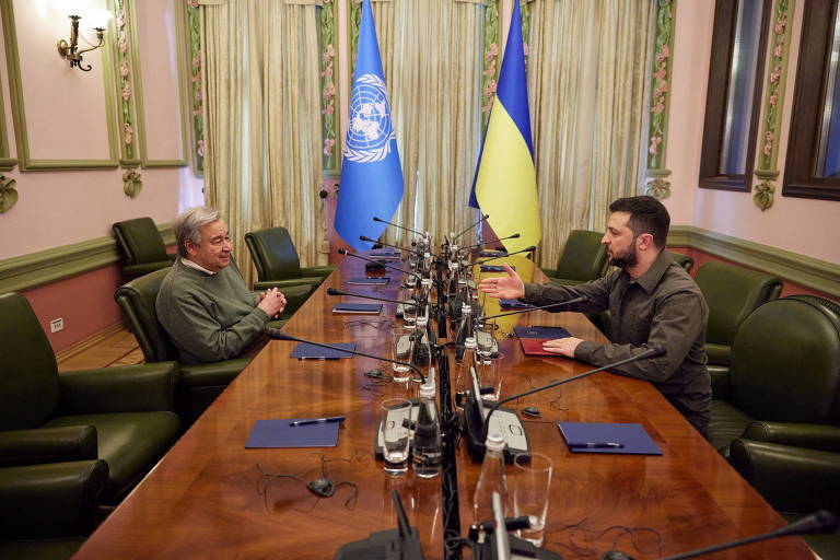 António Guterres e Volodimir Zelenski estão sentados, frente a frente, ao fundo de uma grande mesa de madeira no gabinete do presidente; as bandeiras da ONU e da Ucrânia enfeitam a sala. 