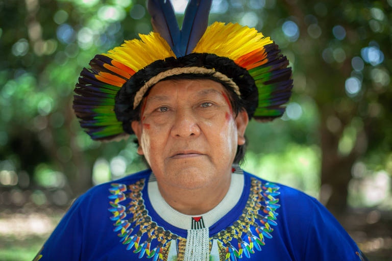 Davi Kopenawa ocupa a maior parte do primeiro plano da foto. Ele é um homem indígena que olha fixamente para a câmera. Usa cocar, uma camisa azul, um colar e tem duas pinturas vermelhas no canto dos olhos. 
